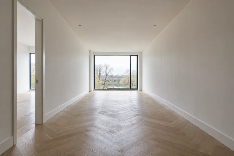 Lichte, lege woonkamer met witte wanden, eiken visgraatvloer en een groot zwart kozijn aan het einde van de ruimte, als sfeerimpressie.